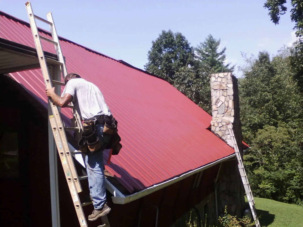 Roofer on a ladder beside a red metal roof and stone chimney — Soffit & Fascia Repair by Roofing Brothers in Bangor